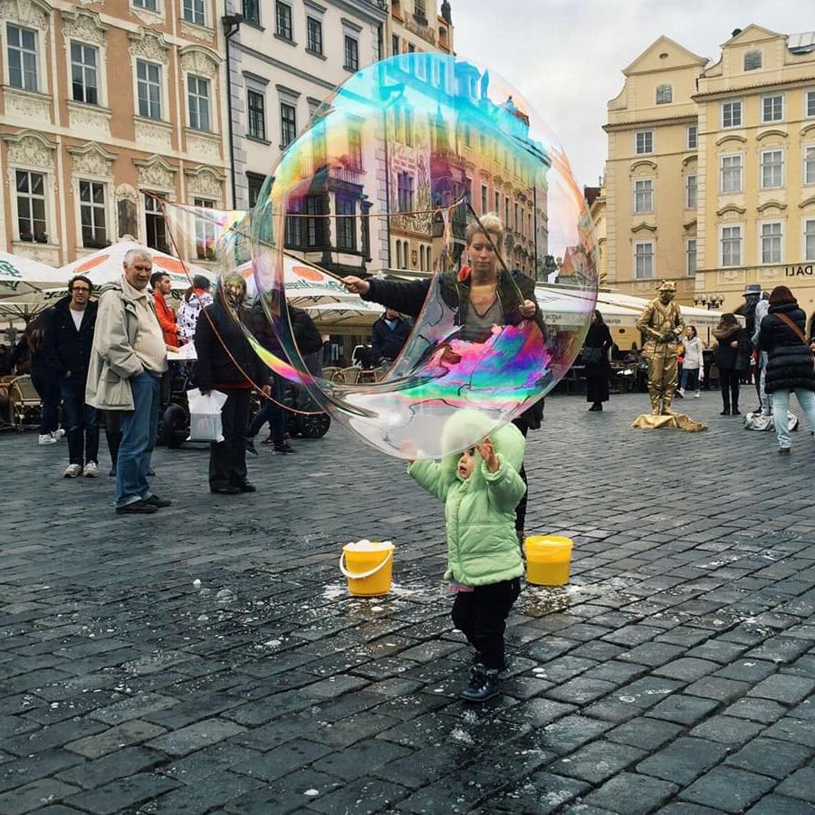 child blows giant rainbow bubble on plaza in Europ